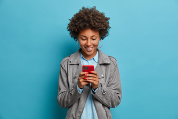 Happy smiling curly haired young woman types message on mobile phone, looks with glad expression at...