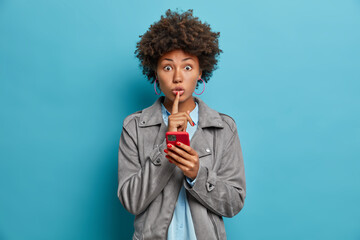 Secret surprised woman with curly hair, dark skin, makes silence gesture, holds mobile phone, creats new profile in social networks, tells secret or confidential information, poses over blue wall - obrazy, fototapety, plakaty