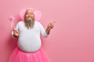 Funny man wears fairy costume, invites you on holiday or costume party, indicates right at blank space, holds magic wand, poses against rosy wall. Dad entertains children during birthday celebration - obrazy, fototapety, plakaty
