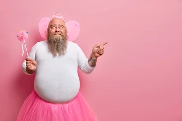 Fotobehang Funny man wears fairy costume, invites you on holiday or costume party, indicates right at blank space, holds magic wand, poses against rosy wall. Dad entertains children during birthday celebration © Wayhome Studio