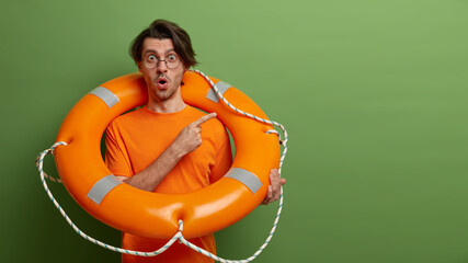 Emotional stunned man poses with safety equipment, uses lifebuoy on rescue, points at copy space, has shocked expression, isolated over green background. Time for swimming. Accident prevention