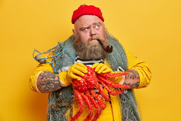 Sailing, sea life and traveling concept. Thoughtful experienced bearded sailor poses with red octopus, smokes pipe travels by ship, isolated on yellow background. Boatswain holds sea creature - obrazy, fototapety, plakaty