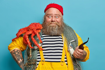 Photo of bearded experienced sailor poses with fishing net, big red crab on shoulders, smoke pipe, welcomes on board, enjoys marine cruise during summer vacation. Bearded captain has sea life