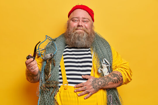Happy pleased sailor keeps hand on big stomach, smokes pipe and enjoys beach party, poses with fishing net, stands indoor in traditional clothing. Succesful fisherman on sea boat has nice day