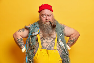 Horizontal shot of plump bearded sailor in overalls keeps hands on hips, thinks deeply while smokes...