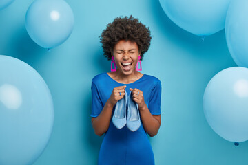 Photo of joyful woman rejoices buying new outfit, holds blue stylish shoes to fit dress, dresses on special occasion, going to celebrate birthday. Blue color prevails. Fashion and clothing concept