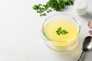 Chicken broth with greens, garlic, onion in glass bowl on white table. Copy space. Close up.