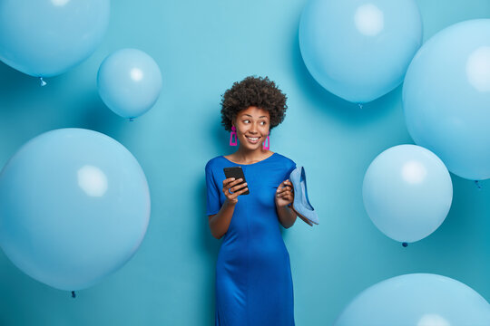 Beautiful lady in blue dress, holds high heel shoes, holds mobile phone, sends invitation for birthday party, surrounded by helium balloons, chooses outfit for special occasion, enjoys event - Powered by Adobe