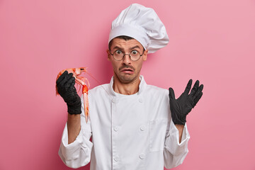 Scared emotional chef holds uncooked crayfish, prepares sea cuisine, healthy vegetarian food, wears white uniform and rubber gloves, hears bad comments from customerrs, isolated on pink background