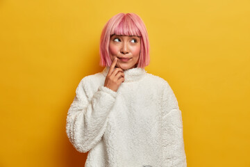 Indoor shot of suspicious thoughtful woman with eastern appearance, makes assumption, thinks about something, keeps finger near mouth, dressed in white jumper, poses indoor, yellow background