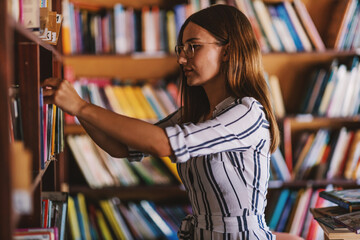 Young gorgeous female student standing next to book shelves and searching for the book for exams.