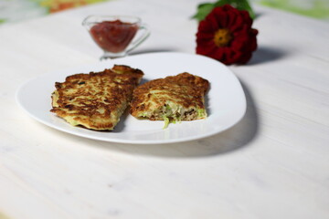 Fried squash pancakes with meat mince and cheese on white plate with tomato sauce and red flower on wooden table