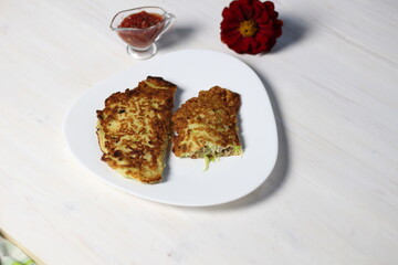Fried squash pancakes with meat mince and cheese on white plate with tomato sauce and red flower on wooden table