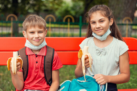 Back To School Concept. Small Girl And Boy Having Lunch Outdoors Sitting On School Bench, Wearing Protective Masks