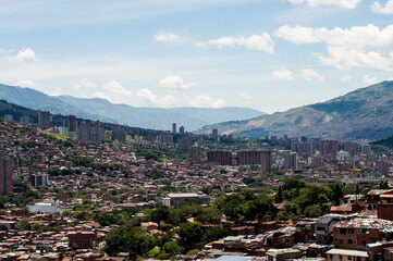 Naklejka premium Sunny hilly urban landscape Medellin city. Buildings and trees in mountains with blue and cloudy sky. Andes mountain range in the background.