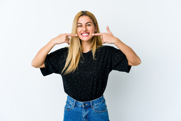 Young blonde woman isolated on white background smiles, pointing fingers at mouth.