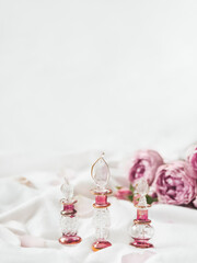 Three graceful bottles for perfume or essential oil on white crumpled fabric. Pink glass bottles with eastern ornament. Pink rose bouquet and petals as decoration.