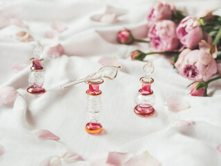 Three graceful bottles for perfume or essential oil on white crumpled fabric. Pink glass bottles with eastern ornament. Pink rose bouquet and petals as decoration.