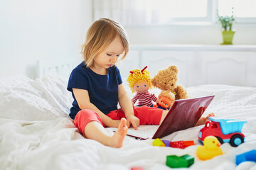 Toddler girl with laptop and toys in bed