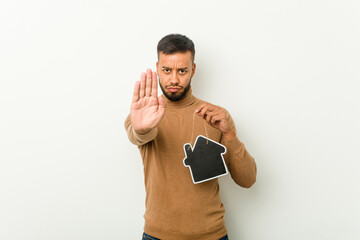 Young south-asian man holding a home icon standing with outstretched hand showing stop sign,...