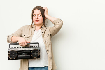 Young curvy woman holding a retro radio touching back of head, thinking and making a choice.