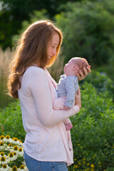 Medium vertical profile shot of pretty smiling red-haired young woman holding her newborn baby girl tenderly while standing in blooming garden