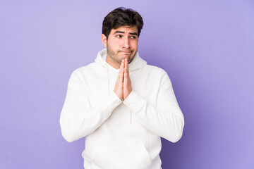 Young man isolated on purple background praying, showing devotion, religious person looking for divine inspiration.
