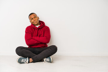 Young latin man sitting on the floor isolated frowning face in displeasure, keeps arms folded.