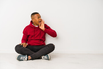 Young latin man sitting on the floor isolated shouting and holding palm near opened mouth.