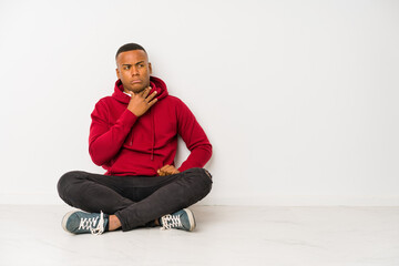Young latin man sitting on the floor isolated suffers pain in throat due a virus or infection.