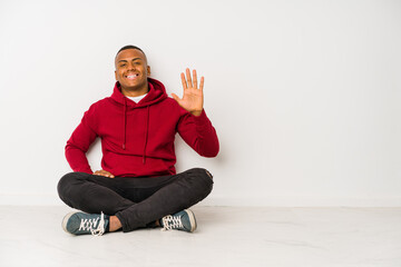 Young latin man sitting on the floor isolated smiling cheerful showing number five with fingers.