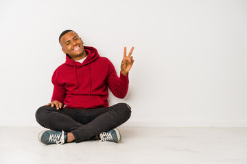 Young latin man sitting on the floor isolated joyful and carefree showing a peace symbol with fingers.