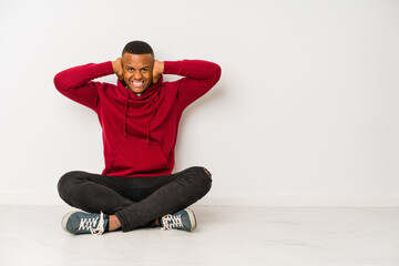 Young latin man sitting on the floor isolated covering ears with hands trying not to hear too loud sound.