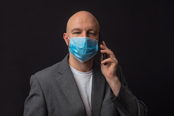 Portrait of bald man in grey suit wearing blue face mask and talking on his smart phone. Dark background. Looking at the camea.
