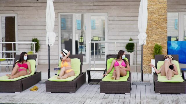 social distance, beautiful young female friends in swimsuits on sun loungers comply with new travel norms and wear protective medical mask to protect against virus and infection during coronavirus