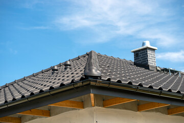 The roof of a single-family house covered with a new ceramic tile in anthracite against the blue sky, visible ridge tile, system chimney and ceramic ventilation fireplace on the roof.