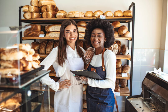 We love our job! Beautiful young women in apron standing in bakery shop. Shot of two women holding a selection of freshly baked breads in their bakery.