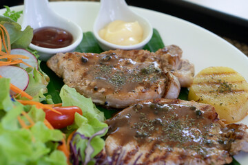 Close up center of food pork steak and a blurry shot of vegetable salad, sauce, and sliced potatoes on a white plate. Ready-to-eat food.