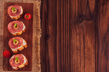 Fototapeta premium Delicious canapes with salami, cream cheese, olives and tomatos on a wooden background - Top view with copy space