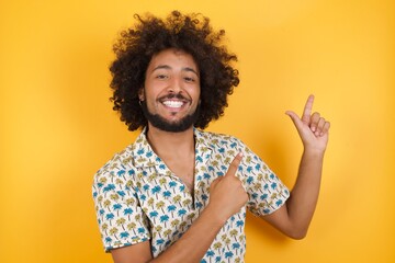 Young man with afro hair over wearing hawaiian shirt standing over yellow background indicating with forefinger empty space showing best low prices