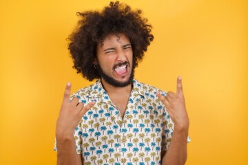 Young man with afro hair over wearing hawaiian shirt standing over yellow background showing rock and roll hand gesture