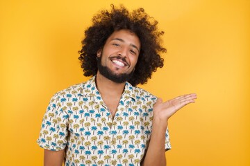 Fototapeta na wymiar Young man with afro hair over wearing hawaiian shirt standing over yellow background smiling cheerful presenting and pointing with palm of hand looking at the camera.