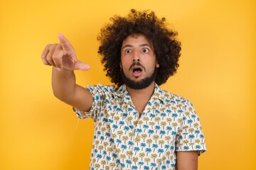 Young man with afro hair over wearing hawaiian shirt standing over yellow background Pointing with finger surprised ahead, open mouth amazed expression, something on the front.