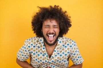 Young man with afro hair over wearing hawaiian shirt standing over yellow background smiling and laughing hard out loud because funny crazy joke with hands on body. - obrazy, fototapety, plakaty