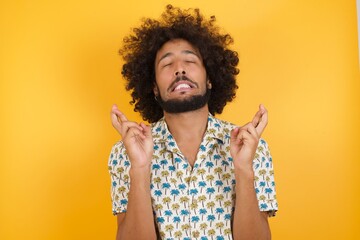 Young man with afro hair over wearing hawaiian shirt standing over yellow background gesturing finger crossed smiling with hope and eyes closed. Luck and superstitious concept.