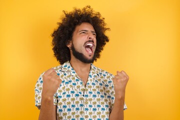 Young man with afro hair wearing hawaiian shirt standing over yellow wall celebrating surprised and amazed for success with arms raised and eyes closed. Winner concept.