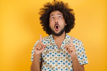 Young man with afro hair wearing hawaiian shirt standing over yellow wall amazed and surprised looking up and pointing with fingers and raised arms.
