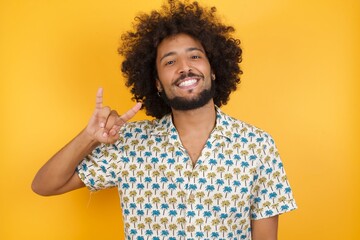 Young man with afro hair wearing hawaiian shirt standing over yellow wall doing a rock gesture and...