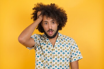 Embarrassed attractive Young man with afro hair wearing hawaiian shirt standing over yellow wall, with shocked expression, expresses great amazement, Puzzled man poses indoor