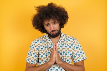 Positive adorable Young man with afro hair wearing hawaiian shirt standing  smiles happily, glad to receive pleasant news from interlocutor, keeps palms together, People emotions concept.